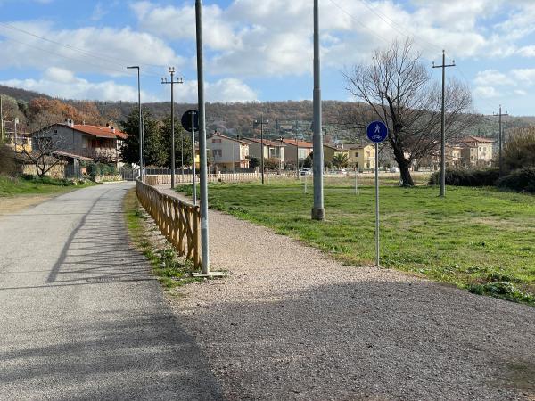 Sortie Monte del Lago : piste cyclable/piétonne plate à fond de gravier. Sur la gauche, une clôture la sépare de la route goudronnée.